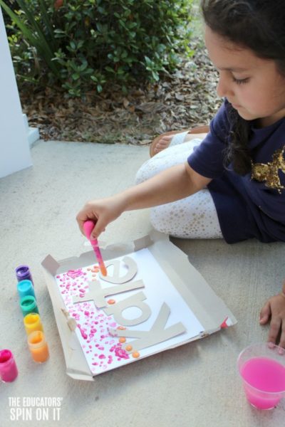 Child drip painting with rainbow colored paints onto paper with stenciled name