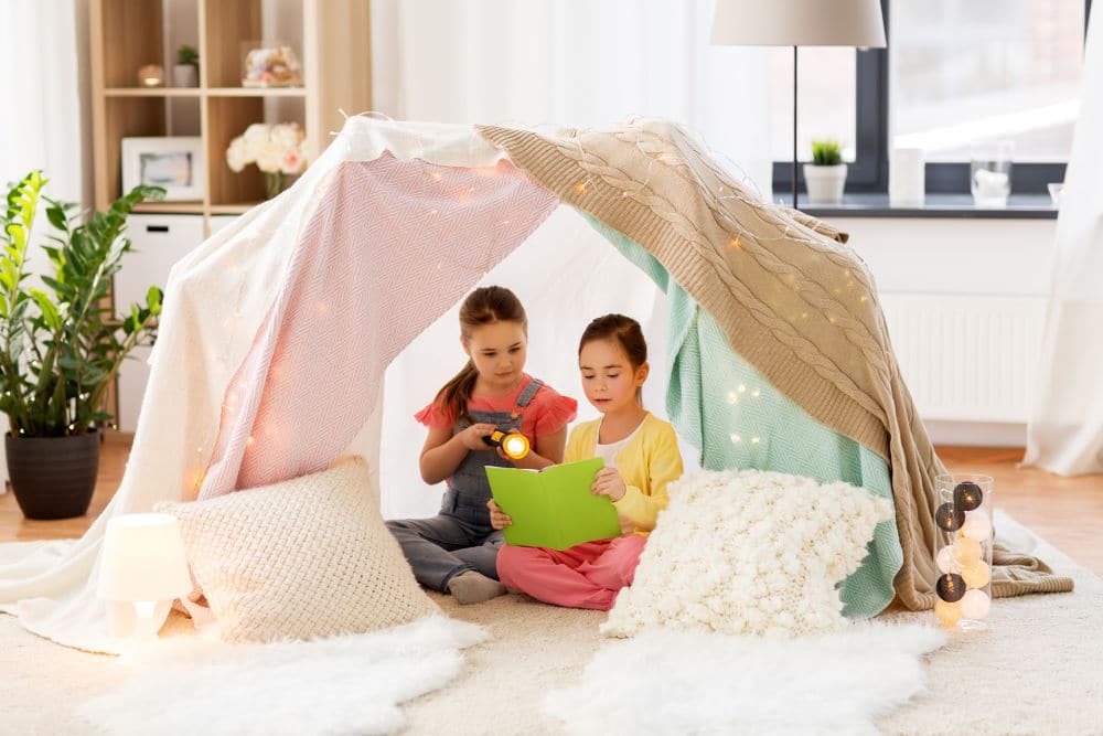 Indoor tent made with blankets and two young girls reading a book
