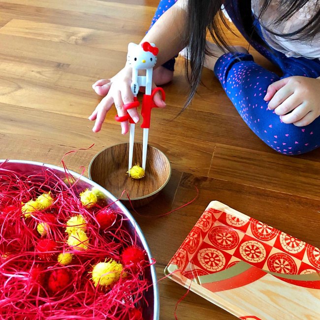 Young girls learning to use chopsticks with red and yellow pom poms in silver bowl