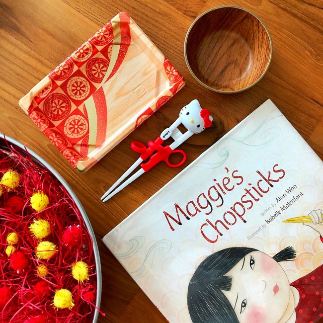 Maggie's Chopsticks book with practice chopsticks and red and yellow pom poms in silver bowl