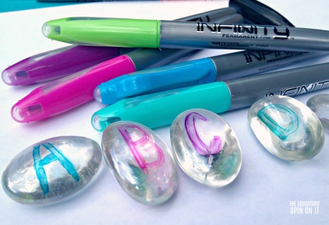 Writing alphabet letters on glass gems with colorful permanent markers for kids reading game.