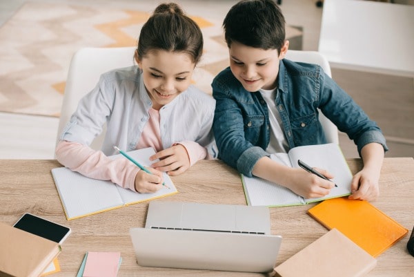 siblings working on school work at home at table with laptop, paper and pencils