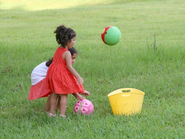 two girls tossing balls into yellow bucket in backyard