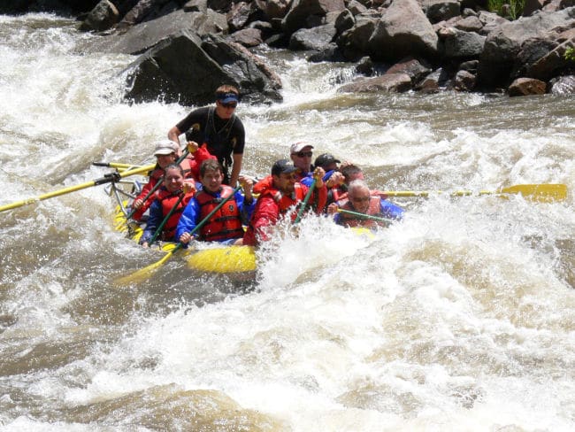 Large Group of people in vests rafting down the Colorado river with waves and rocks in background. 