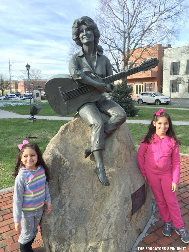 dolly-parton-statue-sevierville-tennessee - The Educators' Spin On It Bronze statue of a woman with a guitar perched on a rock in a park, with two smiling girls standing beside it.
