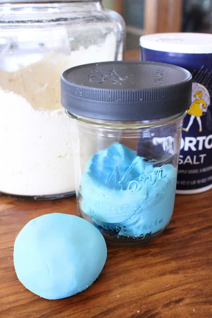 Homemade-playdough-recipe-finished 1 - The Educators' Spin On It Blue bath bomb and a jar of blue bath mixture on a wooden surface, with a salt shaker in the background.