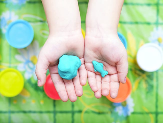 child-holding-playdough - The Educators' Spin On It Two small hands hold blue playdough blobs—a large one in the left palm and a smaller one in the right—on a colorful play area backdrop.