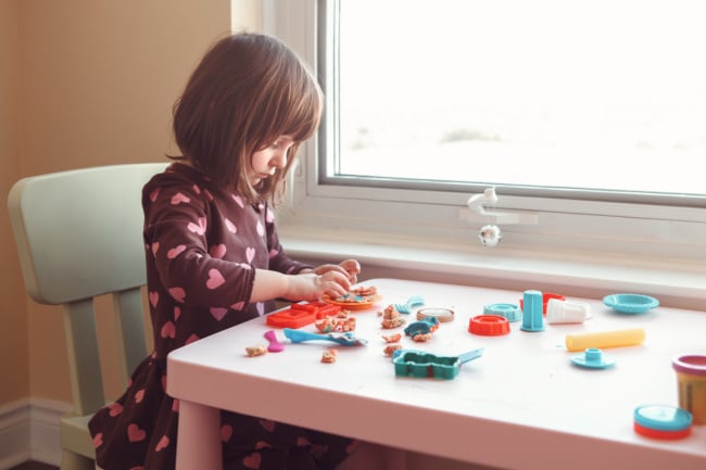 child-playing-with-playdough - The Educators' Spin On It Young girl sits at a white table by a window, molding colorful clay with various tools nearby, focused on her craft project.