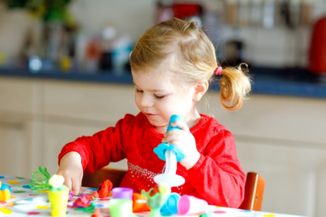 child-using-tools-with-playdough - The Educators' Spin On It Young girl in a red shirt sits at a kitchen table, shaping colorful playdough with tools.