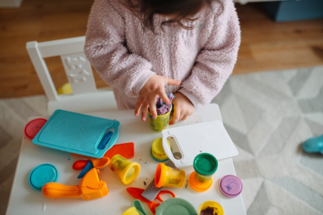 playdough-and-playdough-toys - The Educators' Spin On It Child in a pink fuzzy sweater playing with colorful Play-Doh on a white table, using sculpting tools and containers nearby, at home.